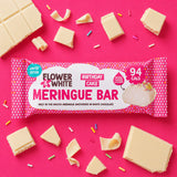 A packaged white chocolate meringue bar wrapped in pink-and-white polka dot paper packaging, surrounded by broken white chocolate pieces and colorful sprinkles on a bright pink background. The package reads 'Flower & White', 'Birthday Cake', and 'Meringue Bar'.