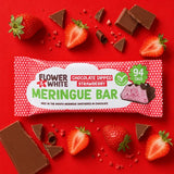 A packaged Flower & White Chocolate Dipped Strawberry Meringue Bar laid on a bright red background with strawberries, chocolate pieces, and crumbs around it.