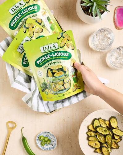 A hand holding a bag of Dj&a Pickle-Licious jalapeño chips, with two more bags in a basket on a light wood table surrounded by small dishes, a green pepper, and a potted plant.