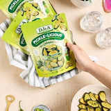 A hand holding a bag of Dj&a Pickle-Licious jalapeño chips, with two more bags in a basket on a light wood table surrounded by small dishes, a green pepper, and a potted plant.