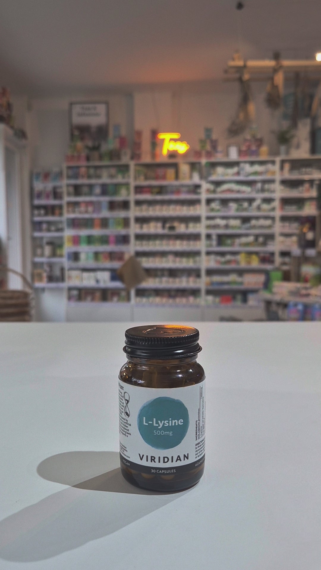 Supplement bottle labeled 'Viridian L-Lysine' on a white surface with a blurred background of shelves.