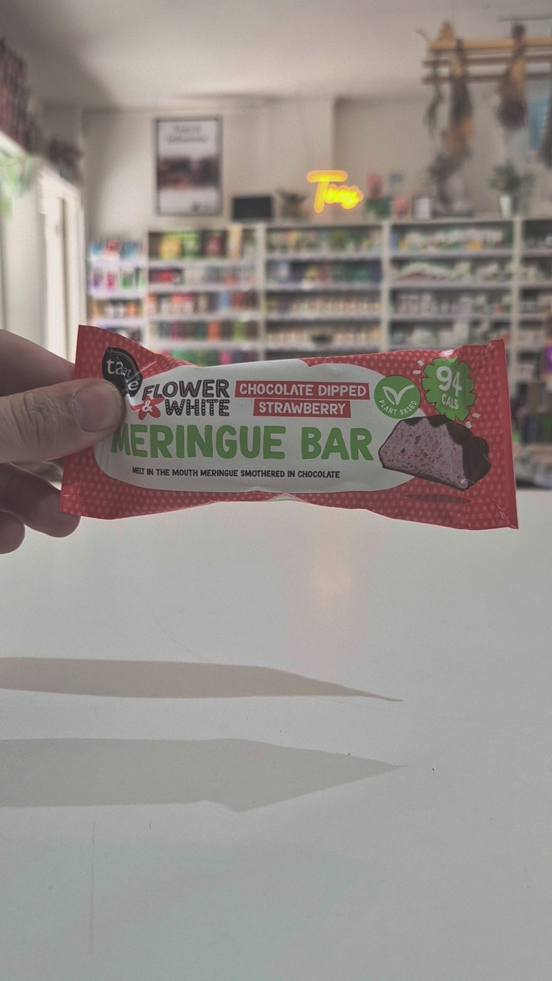 A packaged meringue bar in a red and white wrapper labeled 'Flower & White' and 'Chocolate Dipped Strawberry', placed on a white surface with a blurred store shelf background video