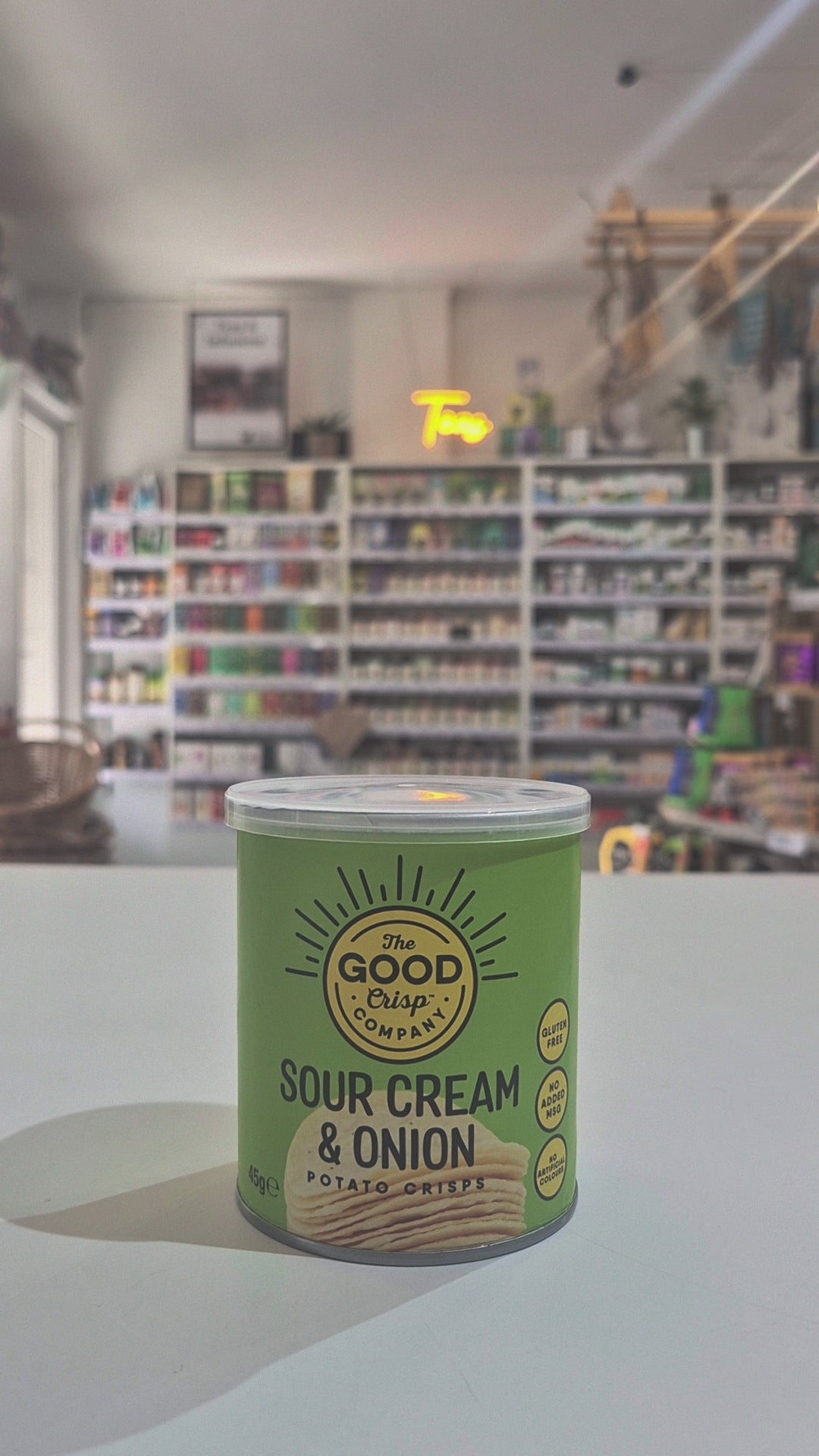Can of 'Sour Cream & Onion' potato crisps from The Good Crisp Company on a white surface with a blurred store background.