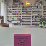 Pink deodorant refill box labeled 'fussy' on a counter with a store background.