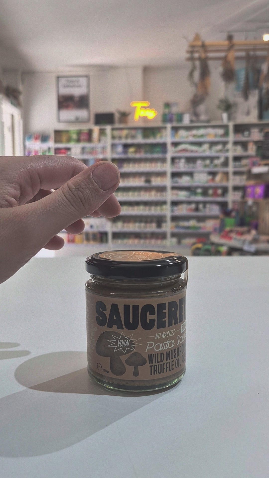 A glass jar of Saucerer Wild Mushroom Truffle Oil pesto with a black lid and brown label, sitting on a white table in a blurred shop interior Video