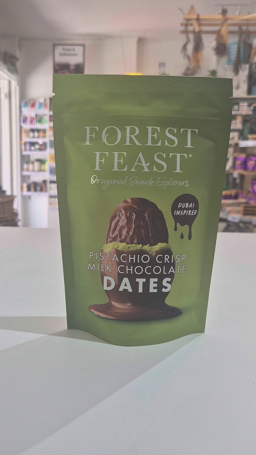 Green package of Forest Feast pistachio crisp milk chocolate dates on a white surface with a store background.