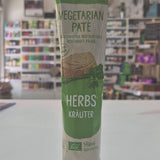 A tube of Tartex Vegetarian Pate with Herbs featuring a green and beige label with a pink cap, shown against a blurred background of a store interior.