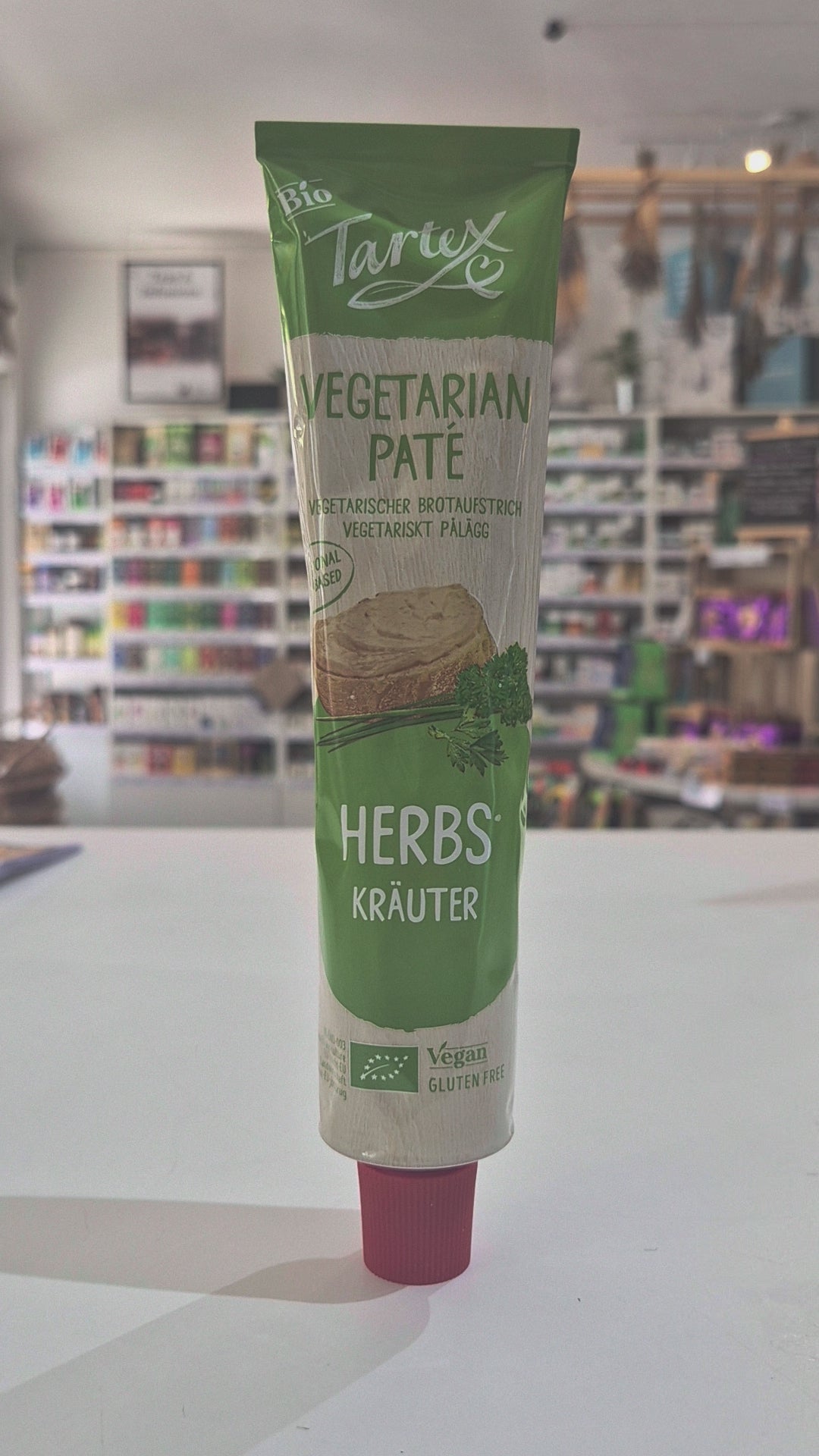 A tube of Tartex Vegetarian Pate with Herbs featuring a green and beige label with a pink cap, shown against a blurred background of a store interior.