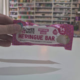A smiling man in a patterned shirt and black apron holds up a packaged Flower & White Meringue Bar in a shop setting with shelves of products behind him.
