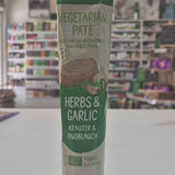 A squeeze tube Tartex Herbs and Garlic with a pink cap, lying horizontally on a white surface with a blurred store interior in the background.