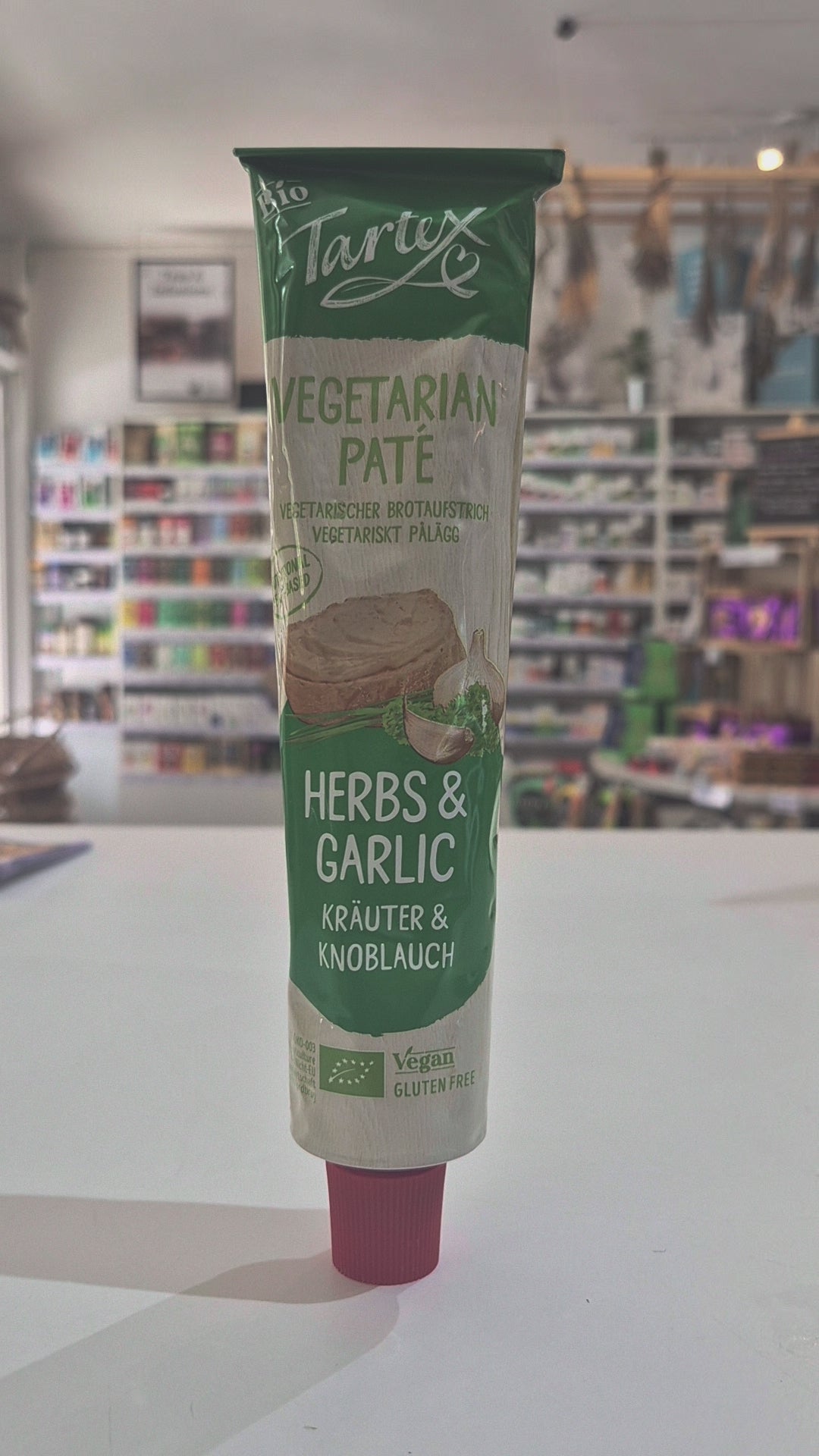 A squeeze tube Tartex Herbs and Garlic with a pink cap, lying horizontally on a white surface with a blurred store interior in the background.