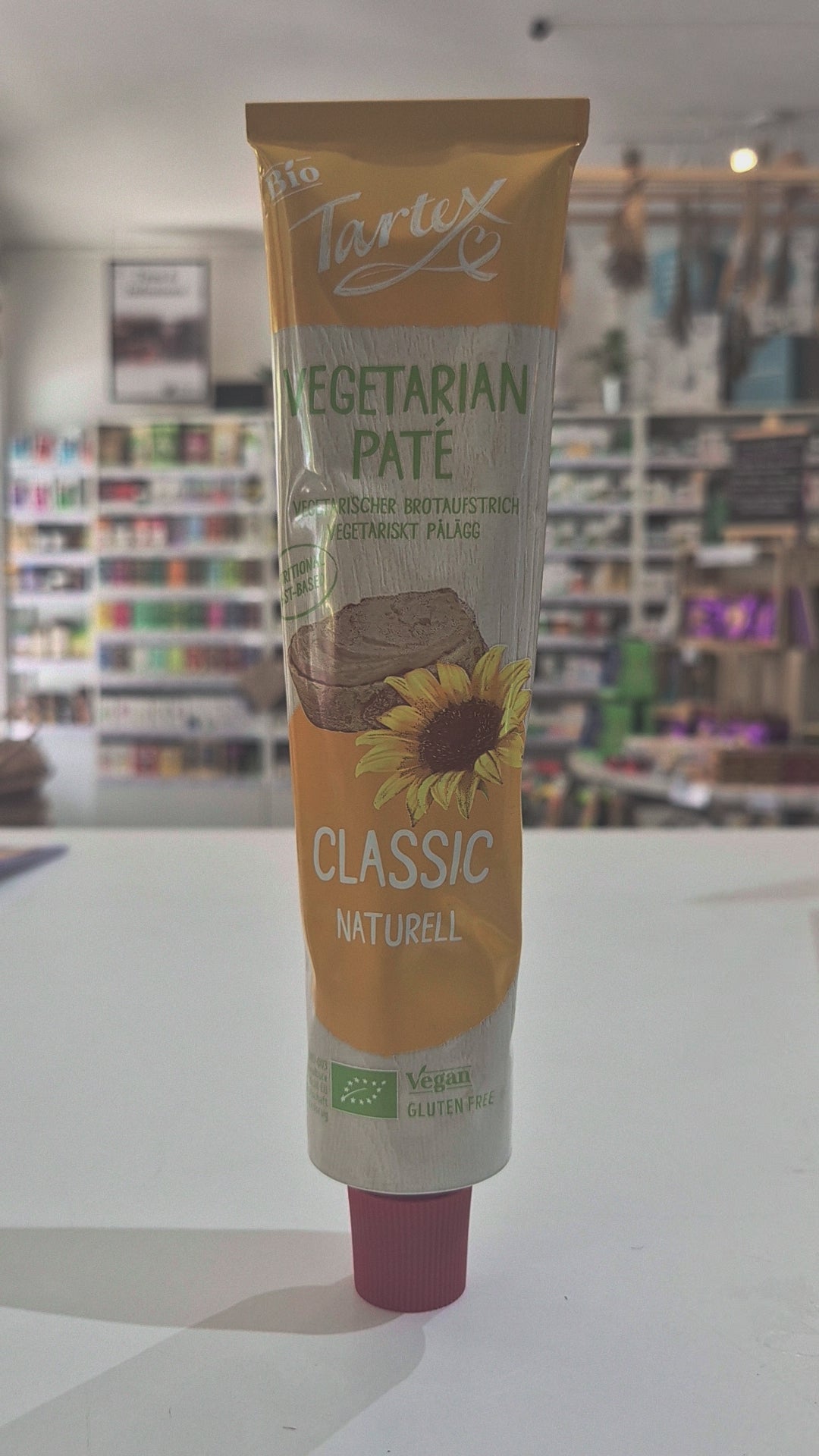 A tube of Tartex Vegetarian Pate Classic spread, featuring a yellow and white label with a sunflower graphic, lying on a white surface with a blurred store background.