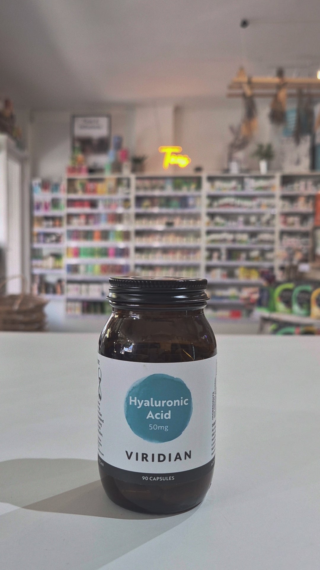 Brown supplement bottle labeled 'Hyaluronic Acid' by Viridian on a white surface with a blurred store background.