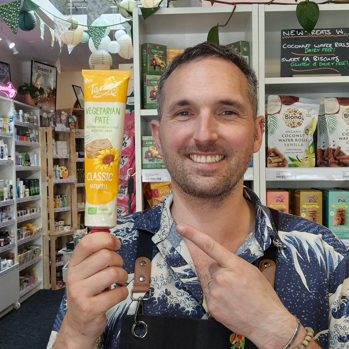 A smiling man wearing a black Health & Remedy apron over a blue and white patterned shirt, posing in front of shelves stocked with various products inside what appears to be a retail shop with Tartex Classic Pate