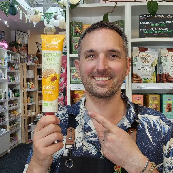 A smiling man wearing a black Health & Remedy apron over a blue and white patterned shirt, posing in front of shelves stocked with various products inside what appears to be a retail shop with Tartex Classic Pate