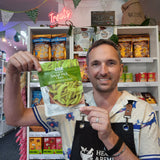 A smiling man in a shop holds up a bag of DJ&A Snaps Delicious Organic Snap Peas, pointing at the green peas on the packaging. The background shows shelves stocked with various snack products.