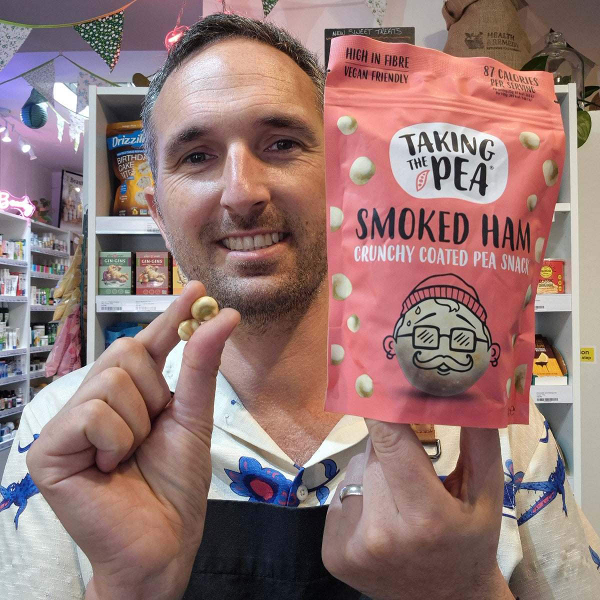 A smiling man in a shop holds up a pink bag of 'Taking the Pea' Smoked Ham crunchy coated pea snacks next to his fingers, which also hold a pea snack.