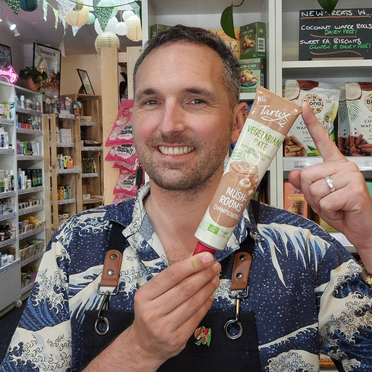 A smiling man in a navy and white wave-patterned shirt and black apron holds up a tube of Tartex Vegetarian Yeast Free Mushroom pate in a shop setting.