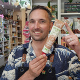 A smiling man in a navy and white wave-patterned shirt and black apron holds up a tube of Tartex Vegetarian Yeast Free Mushroom pate in a shop setting.