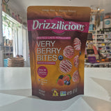 A stand-up pouch of Drizzilicious Very Berry Bites snack mix placed on a white countertop in a retail store setting. The package is brown and orange with a pink brand banner and berry illustrations.