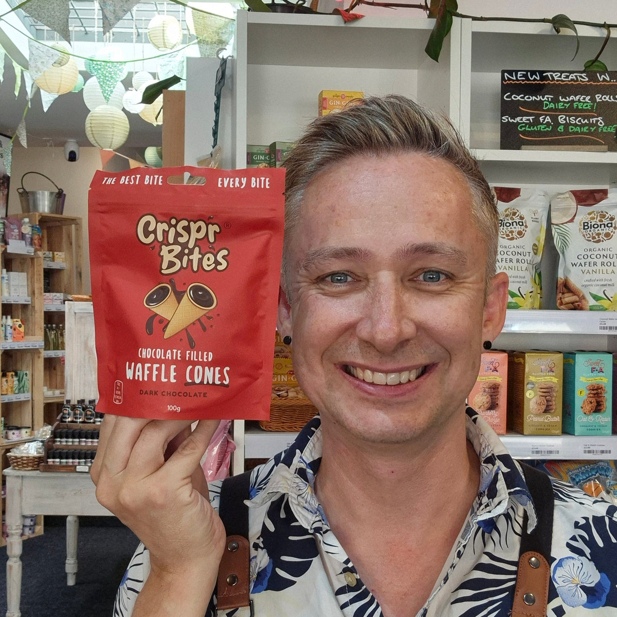 A smiling man in a patterned shirt and black Health & Remedy apron holds up a red bag of Crispr Bites Chocolate Flavour Waffle Cones in a shop setting.