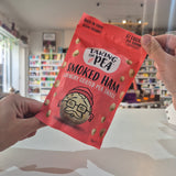 A person holds a red bag of Taking the Pea Smoked Ham crunchy coated pea snack in a shop setting with shelves blurred in the background.