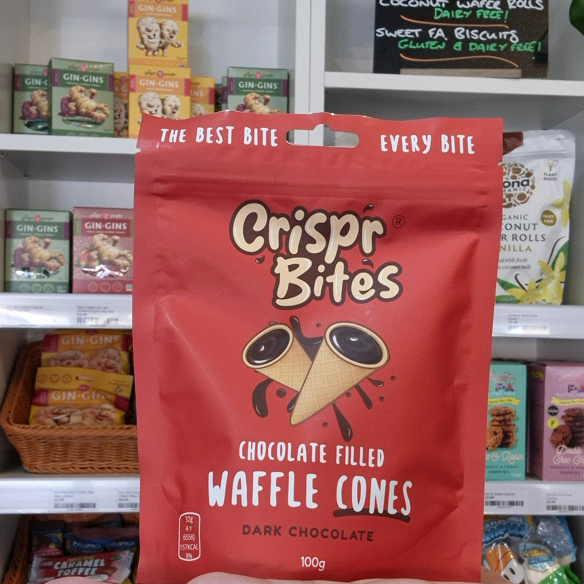 A hand holding a red bag of Crispr Bites Chocolate Filled Waffle Cones in front of a white shelf filled with various packaged snacks.