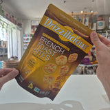 A person holds a bag of Drizzilicious French Toast Bites in a shop setting. The packaging is brown and gold with images of the toast bites and text reading 'French Toast Bites' and 'Mini Rice Cakes Reinaparen!'.