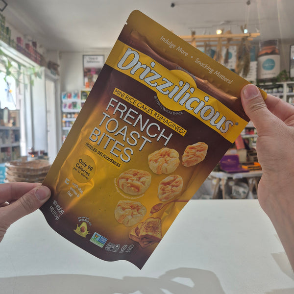 A person holds a bag of Drizzilicious French Toast Bites in a shop setting. The packaging is brown and gold with images of the toast bites and text reading 'French Toast Bites' and 'Mini Rice Cakes Reinaparen!'.