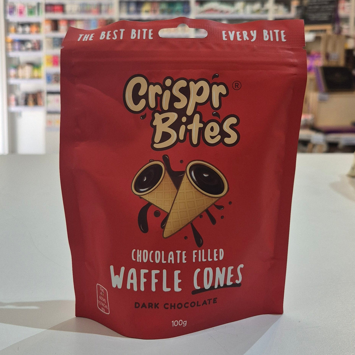 A red stand-up pouch of Crispr Bites Chocolate Filled Waffle Cones placed on a white countertop in a blurred retail shop setting.