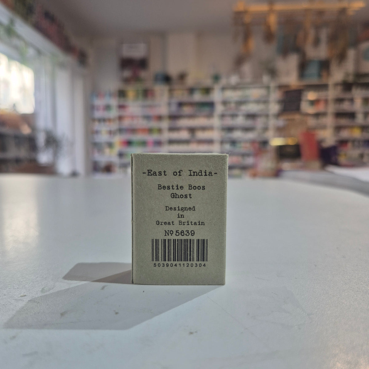 A small square product box standing upright on a white tabletop in a blurred shop interior. The box is light gray with black text reading “-East of India-” and “Beatie Boos Ghost,” plus a barcode and “Designed in Great Britain.