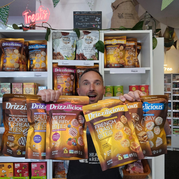 A man in a retail store holds up multiple bags of Drizzlicious snack bites in front of white shelves stocked with other snack products. The bags shown include Cookie & Cream Bites, Salty Caramel Bites, Very Berry Bites, and French Toast Bites.