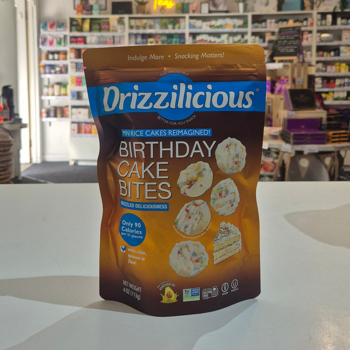 A bag of Drizzilicious Birthday Cake Bites snack stands on a white counter in a retail store. The packaging is brown and blue with images of cake pieces.