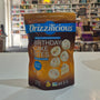 A bag of Drizzilicious Birthday Cake Bites snack stands on a white counter in a retail store. The packaging is brown and blue with images of cake pieces.