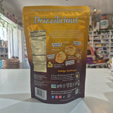 A stand-up pouch of Drizzlicious dried shrimp snacks photographed on a countertop in a retail store. The back of the package is mostly dark brown with a large yellow upper section, nutrition facts, product imagery, and flavor callouts including avocado crunch, chip & sunflower butter, and wholegrain.
