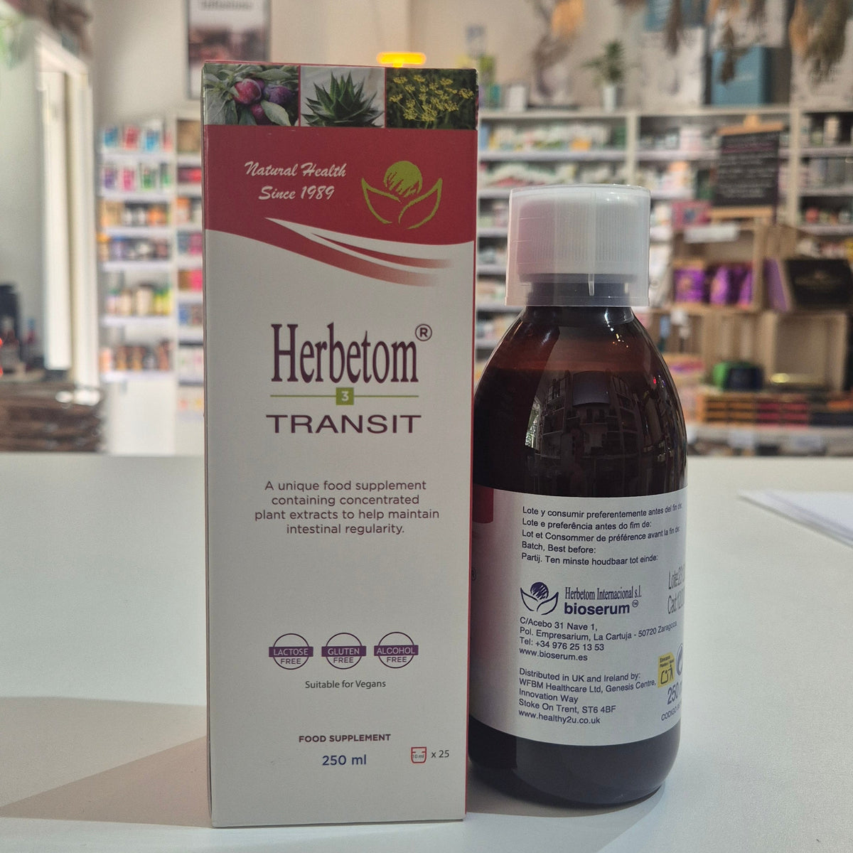 A bottle of Herbetom Transit food supplement next to its retail box, displayed on a white surface with a blurred store background.