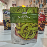 A bag of DJ&A Sugar Snap Pea Crisps in the green houseplant flavor, standing on a white countertop with two other snack bags blurred in the background.