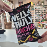 A bag of Well & Truly Crunchies Crunchy Pickles snack chips sits on a white counter in a retail store setting with shelves of products in the background.