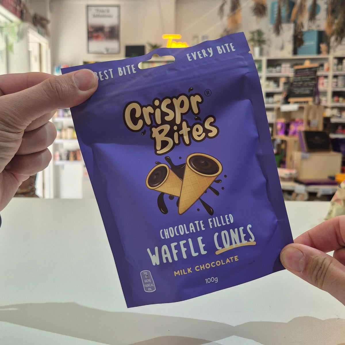 A person holds a purple bag of Crispr Bites Chocolate Filled Waffle Cones in a shop setting.