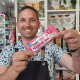 A smiling man in a patterned shirt and black apron holds up a packaged Flower & White Meringue Bar in a shop setting with shelves of products behind him.