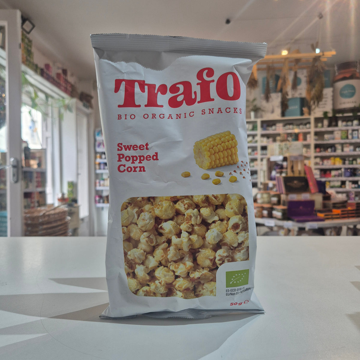 A bag of Trafo Sweet Popped Corn organic snacks standing on a white counter in a retail store. The white bag features red branding, a picture of corn, and a clear window showing the popcorn inside.