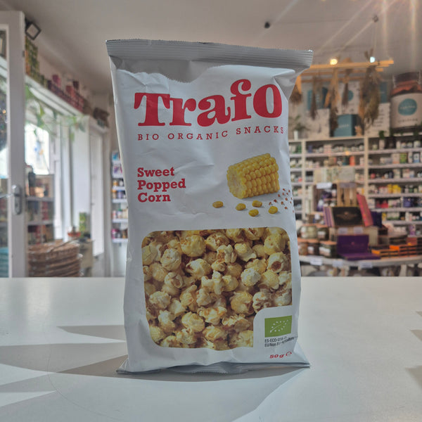 A bag of Trafo Sweet Popped Corn organic snacks standing on a white counter in a retail store. The white bag features red branding, a picture of corn, and a clear window showing the popcorn inside.
