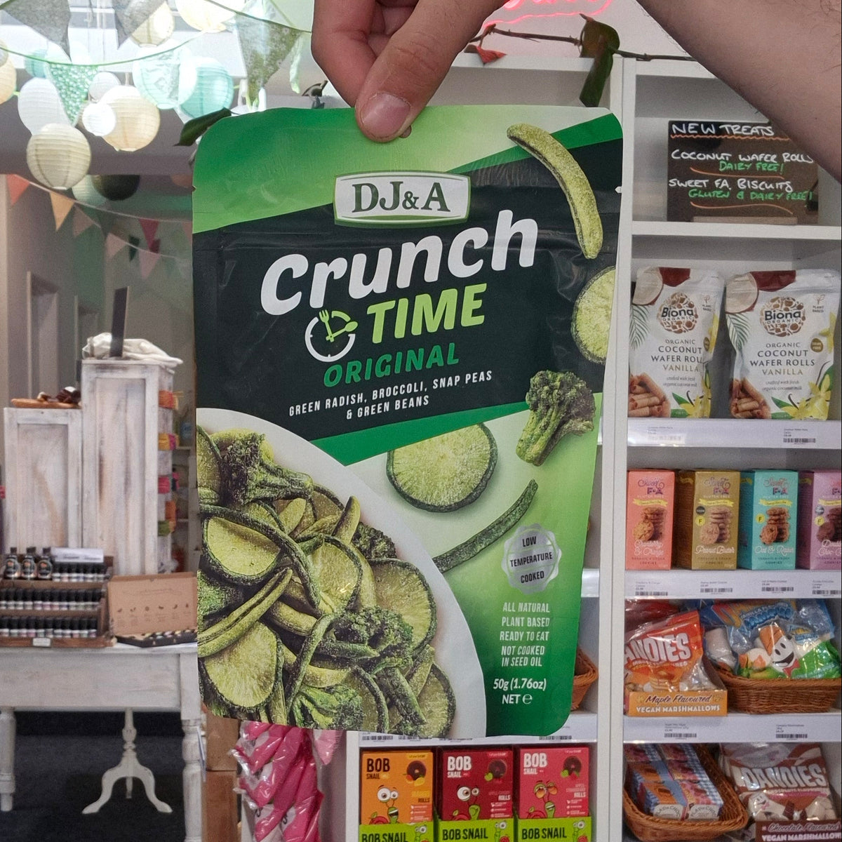 A hand holds a bag of Dj&A Crunch & Time Originals in front of a white retail display shelf filled with packaged snacks inside a store.
