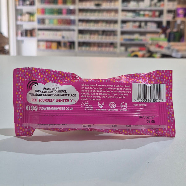 The back of a pink and purple patterned Flower & White product pouch, showing text, a barcode, and icons, photographed on a white surface with a blurred store background.