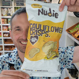 Person holding a package of Nudie Naturals cauliflower crisps in a kitchen setting.