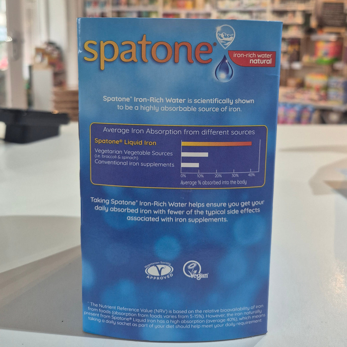 Spatone vitamin supplement packaging held in front of a blurred store interior.