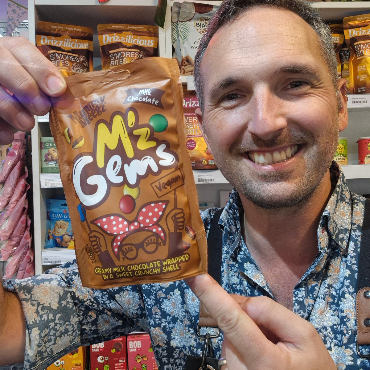 A smiling man in a blue floral shirt holds up a bag of M'z Gems Veggies chocolate chocolate wrapped in a retail store setting.