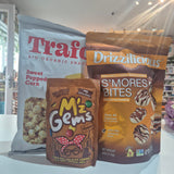 Three snack products displayed together on a countertop: a large bag of Trafo Sweet Popped Corn, a bag of Drizzilicious S'mores Bites, and a bag of M'z Gems. The background shows a retail store interior with shelves and hanging lights.