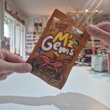 A person holds a bag of Nutty's Mummy Meegz M'z Gems Vegan chocolate wrapped candies in a brightly lit shop setting.
