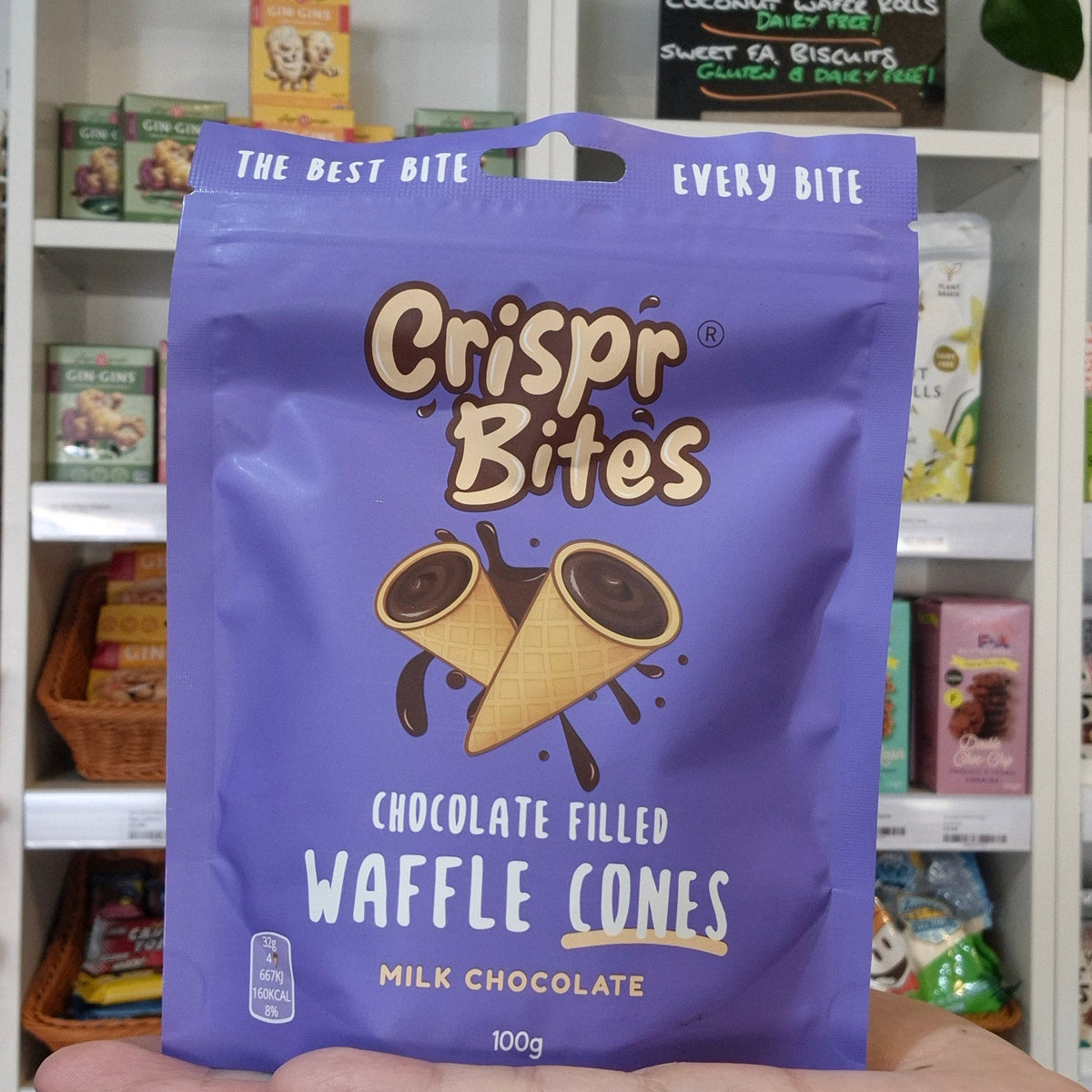 A hand holds a purple bag of Crispr Bites Chocolate Filled Waffle Cones in front of a white shelf filled with packaged snacks and treats.